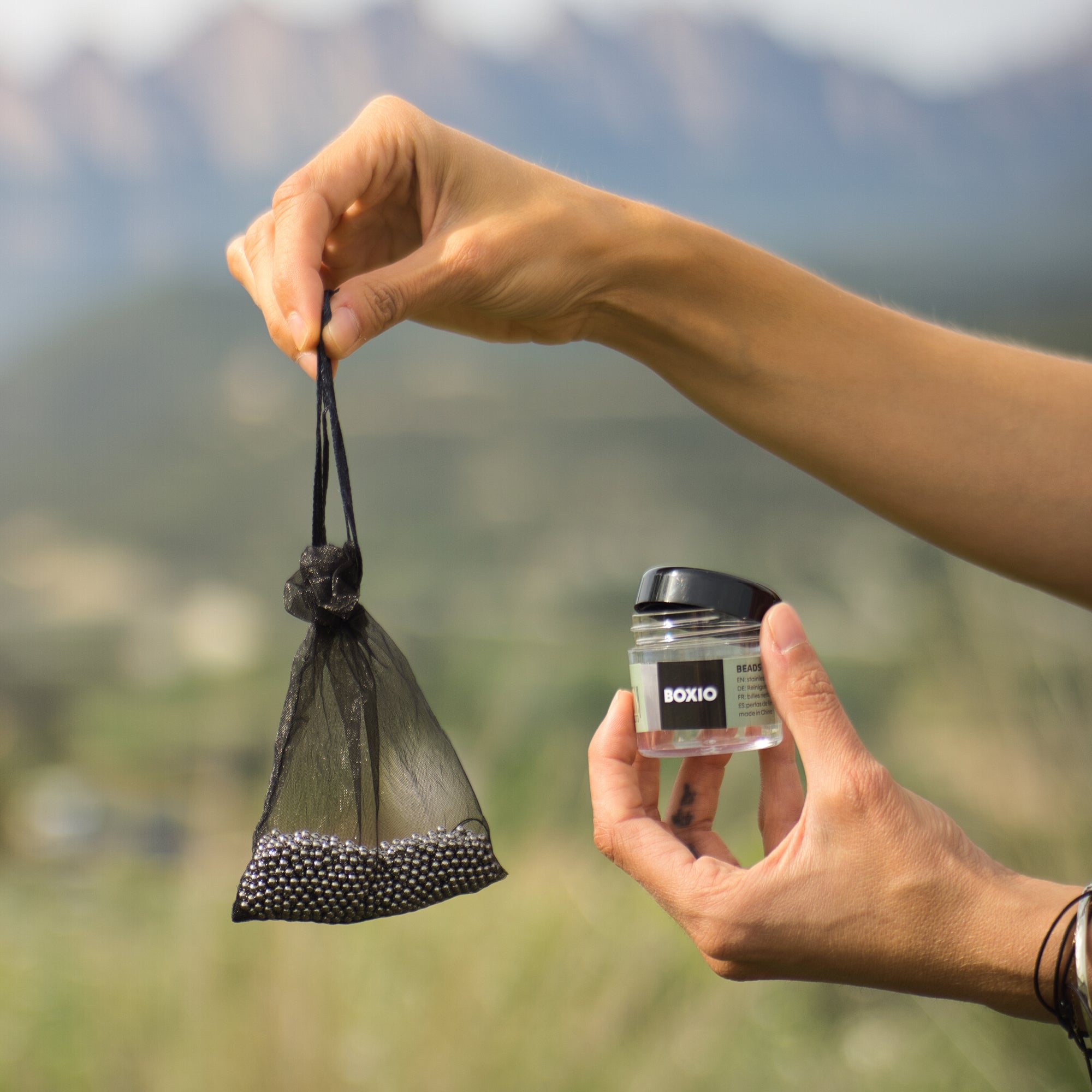 Eine Hand hält einen schwarzen Säckchen mit Perlen und ein Transparentglas mit der Aufschrift "BOXIO" im Freien.