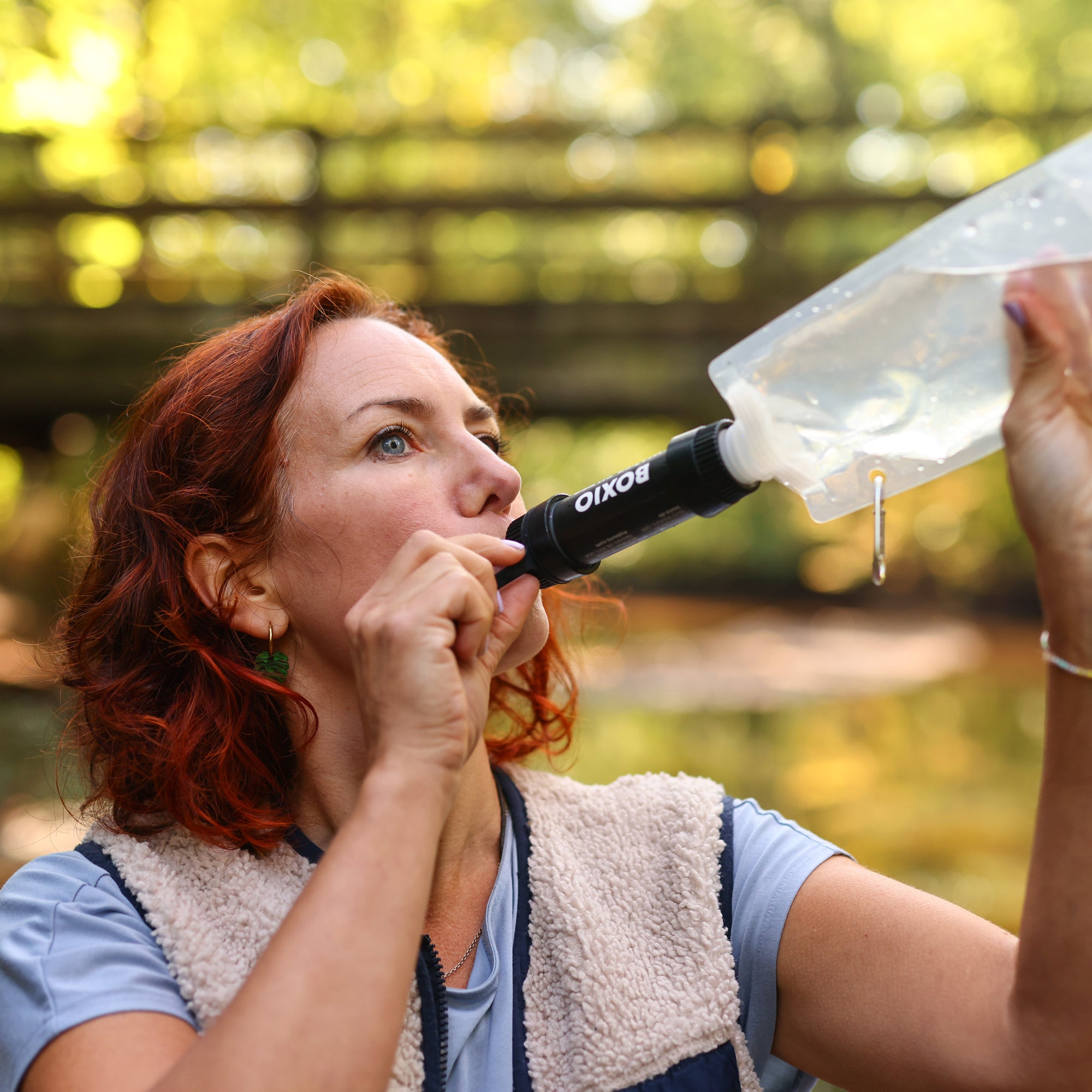 Frau mit rotem Haar trinkt aus einer Wasserflasche mit einem speziellen Trinkaufsatz in der Natur.
