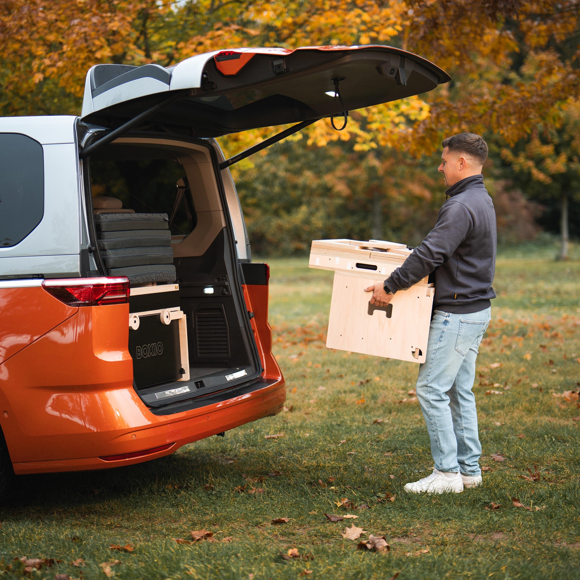 Mann trägt eine Holzbox aus einem offenen, orangefarbenen Van auf eine Wiese mit herbstlichen Bäumen.