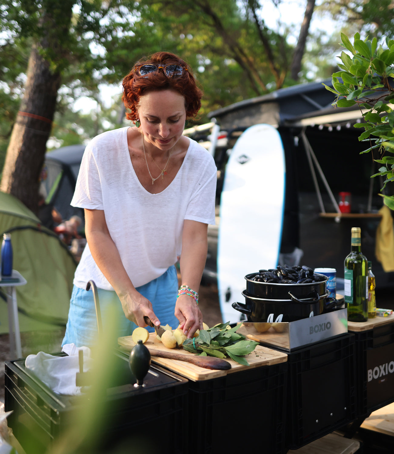 Frau mit roten Locken schneidet Gemüse auf einem Holztisch beim Camping, im Hintergrund Zelt und Kochgeschirr.