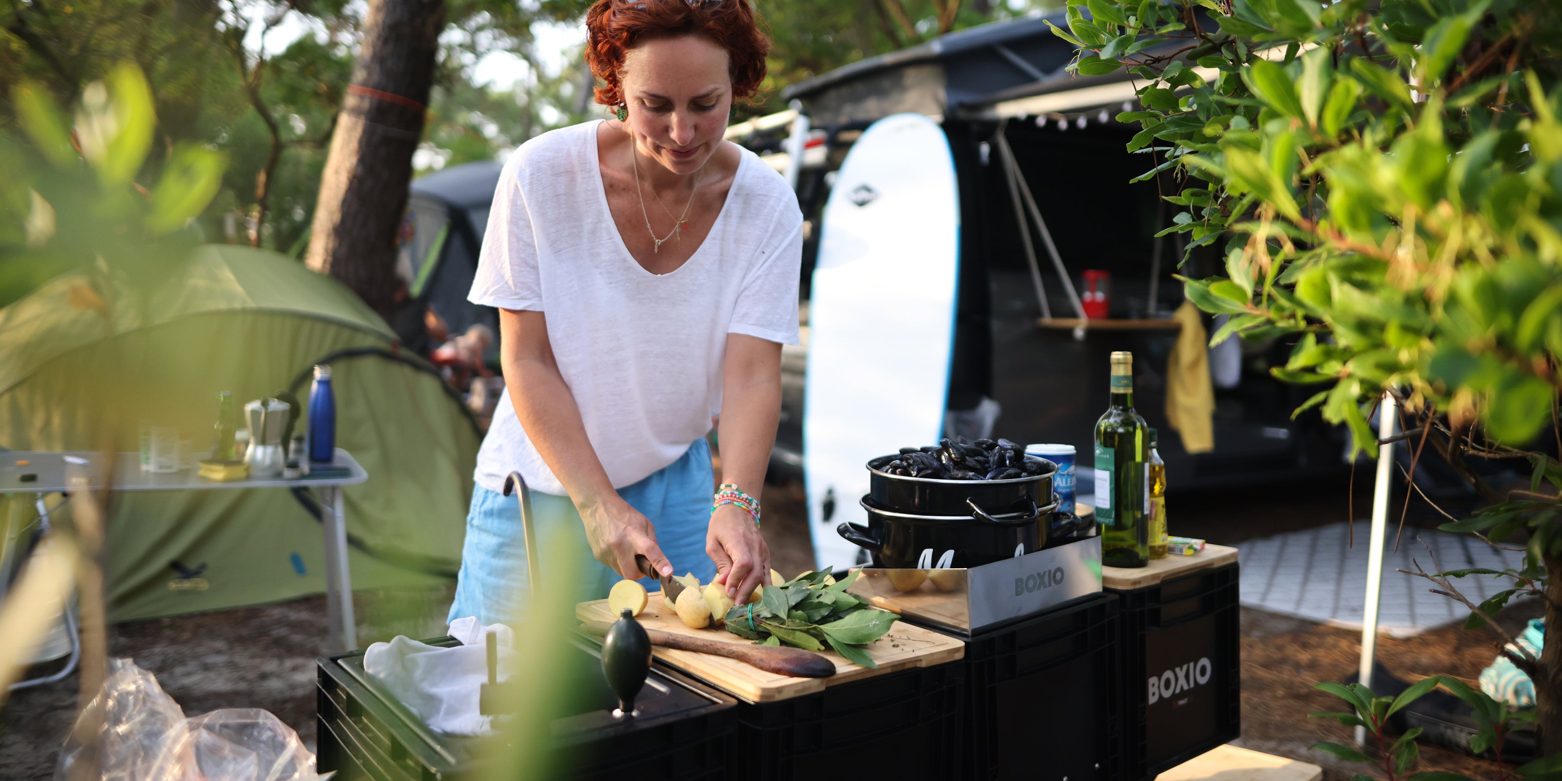 Frau schneidet Gemüse auf einem Holztisch beim Camping, umgeben von Zelten und Kochutensilien.
