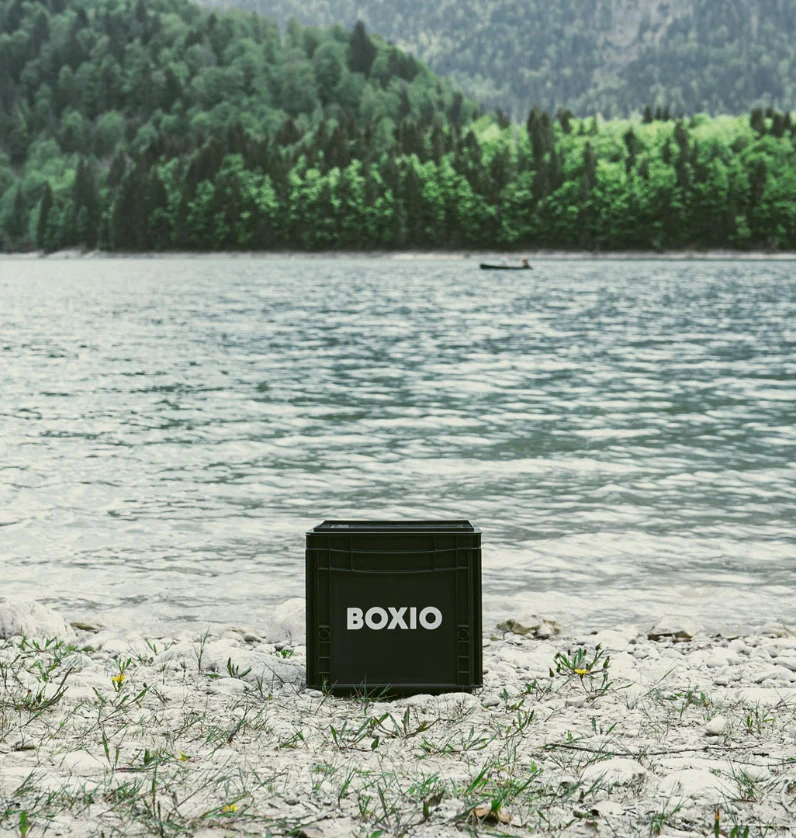 Schwarze BOXIO-Transportbox steht am Ufer eines klaren Gewässers, umgeben von grünem Uferbewuchs und Bergen.